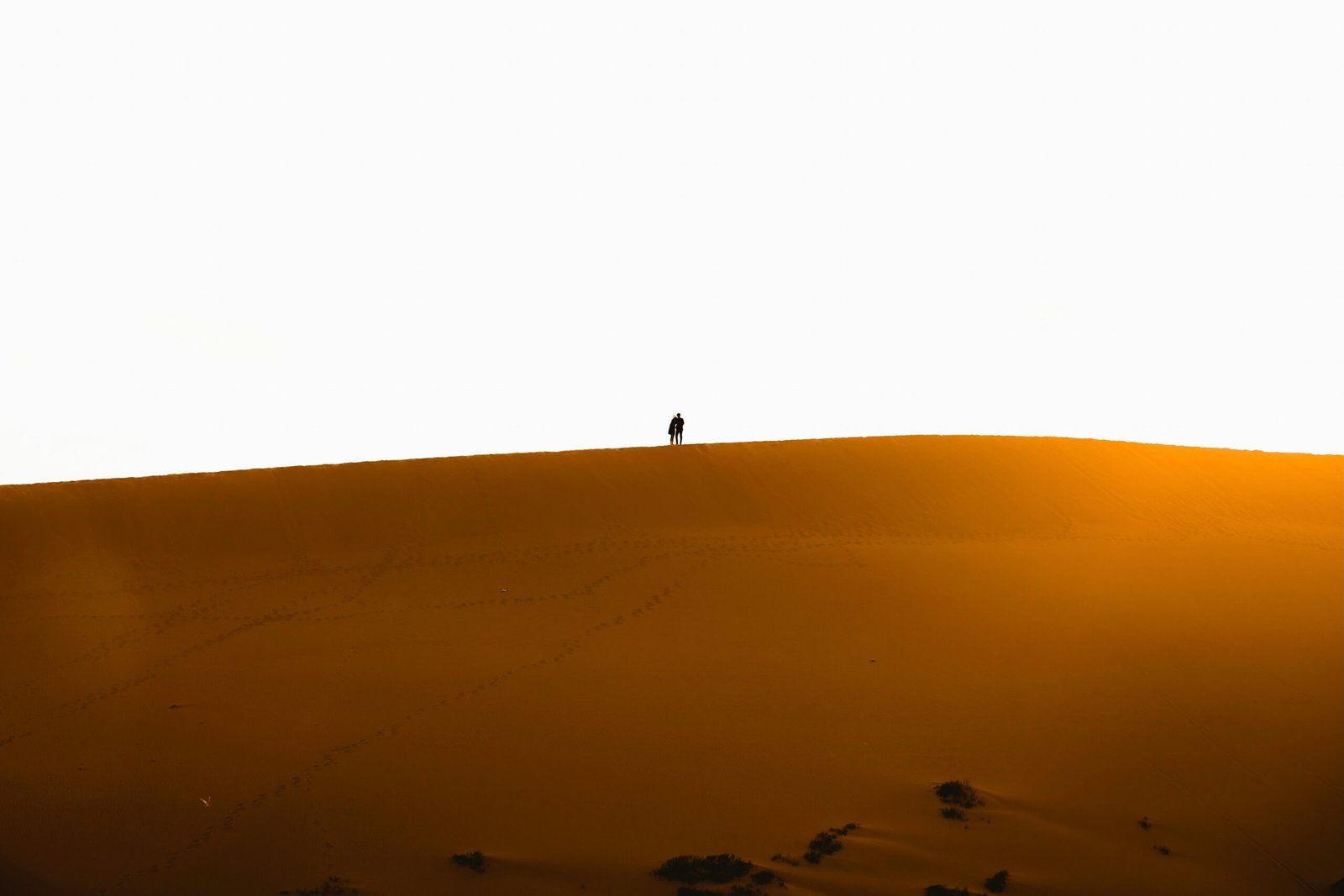 A victorious climber standing on top of a large sand dune with arms raised against a sunset backdrop.