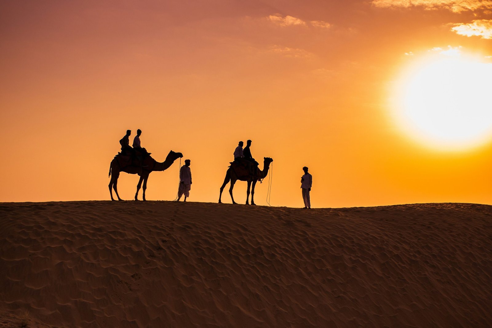 A line of camels crossing vast desert dunes against a fiery orange sky
